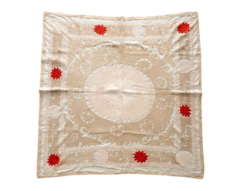 May include: A square, beige textile with intricate embroidered patterns and a central circular design. The textile features red floral accents and a decorative border. The overall design is symmetrical and ornate, suitable for home decor.
