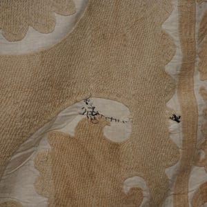 May include: Close-up of a beige textile with intricate patterns and textures. The design features floral and geometric shapes in varying shades of beige and cream, with visible stitching and repairs. The fabric has a slightly worn appearance.