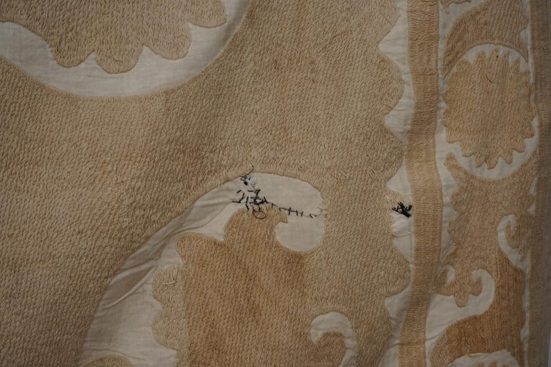 May include: Close-up of a beige textile with intricate patterns and textures. The design features floral and geometric shapes in varying shades of beige and cream, with visible stitching and repairs. The fabric has a slightly worn appearance.