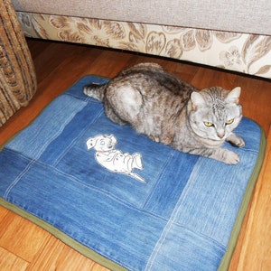 Puede incluir: Un gato atigrado gris descansa sobre una cama cuadrada para mascotas de mezclilla. La cama presenta un diseño de patchwork con un borde verde y una ilustración de dibujos animados de dálmata blanco. La cama está colocada sobre un suelo de madera.