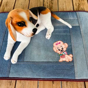 May include: A plush beagle dog toy with orange, white, and black markings lies on a denim pet bed. A small embroidered puppy with a pink bow and basket is also on the bed. The bed has a red border.