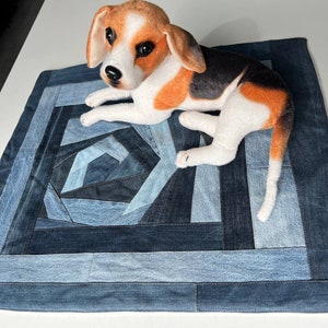 May include: A blue denim dog bed with a geometric pattern made from recycled denim. A small stuffed dog is lying on the bed.