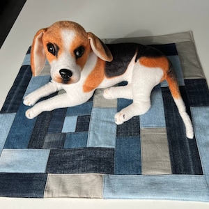 Puede incluir: Un juguete de peluche de beagle acostado en una cama para perros de mezclilla patchwork. La cama está hecha de varios tonos de mezclilla azul y tela gris.