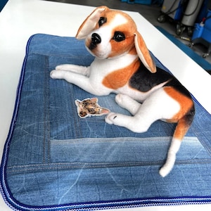 May include: A plush beagle dog toy laying on a blue denim dog bed with a small cat face patch.