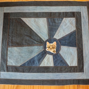 May include: A denim patchwork throw pillow with a circular applique of a cat's face in the center. The pillow has a light blue, dark blue, and black denim patchwork design.