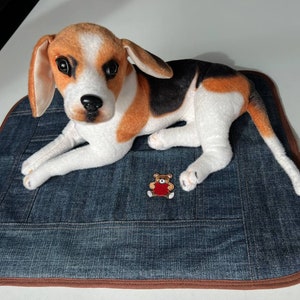 May include: A plush beagle dog toy laying on a blue denim dog bed with a brown border. The dog toy has brown, white, and black fur. A small red embroidered teddy bear is on the bed.