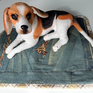 May include: A plush beagle dog toy laying on a blue denim dog bed with a brown and white plaid border.