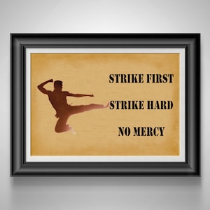 May include: Framed artwork with a black frame. The artwork features a brown silhouette of a person performing a martial arts kick on a tan background. Text reads "STRIKE FIRST, STRIKE HARD, NO MERCY."