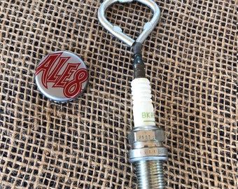 May include: A handmade bottle opener crafted from a spark plug, featuring a silver metal loop handle. The spark plug has the text "BKR" and "U6GT" printed on it. A bottle cap with the word "ALES" is next to the opener.