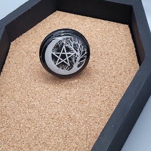 May include: A black resin ring with a white crescent moon, pentagram, and tree design.