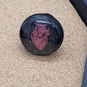 May include: A black round resin cabochon with a red anatomical heart image.