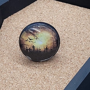 May include: A round glass cabochon with a black metal setting features a vintage-style image of a graveyard at sunset with bats flying overhead. The image is set against a yellow and orange sky with a dark silhouette of trees and tombstones.