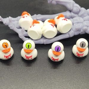 May include: A collection of Halloween-themed rubber ducks. The white ducks have orange beaks, and various colored eyeballs. Some ducks have orange paint on their heads, and red paint dripping down their chests. A gray skeleton hand is in the background.