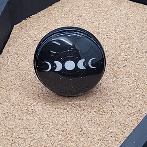 May include: A black round resin cabochon with a white moon phase design. The cabochon is set in a clear resin dome.