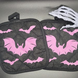 May include: Two black quilted pot holders with pink bat silhouettes. The pot holders are quilted and have a loop for hanging.