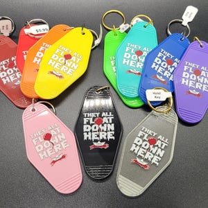 Könnte beinhalten: Eine Sammlung von Schlüsselanhängern im Motel-Stil in verschiedenen Farben, darunter Rot, Orange, Gelb, Grün, Türkis, Blau, Rosa, Schwarz und Grau. Jeder Schlüsselanhänger zeigt den Schriftzug "THEY ALL FLOAT DOWN HERE" mit einem roten Ballon und einem Papierboot.