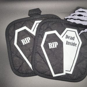 Puede incluir: Dos agarraderas de olla acolchadas negras con contornos blancos de ataúdes. Un ataúd dice "RIP" y el otro dice "Dead Inside".