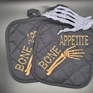 May include: Two black quilted pot holders with a skeleton hand and bone graphic and the text "BONE APPETITE".