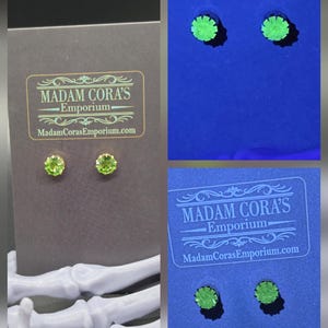 May include: A pair of round, faceted lime green stud earrings displayed on a grey card labeled Madam Cora's Emporium. The earrings exhibit a bright neon glow under ultraviolet light, appearing as vibrant green circles against a blue background.