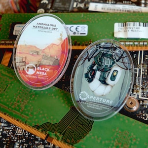 May include: Two oval-shaped clear acrylic pins. One pin features a desert landscape with text that reads "ANOMALOUS MATERIALS DPT. NEW MEXICO" and "BLACK MESA RESEARCH FACILITY." The other pin has a robot graphic and text that reads "A TRUSTED FRIEND IN SCIENCE" and "APERTURE LABORATORIES GLADOS DEVELOPMENT DEPARTMENT."