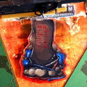 May include: A clear acrylic keychain featuring a dark, tombstone-shaped object with the word HATE printed in red text. The base is surrounded by gray rocks and blue cables, with a black metal lobster clasp attached to the top of the charm.