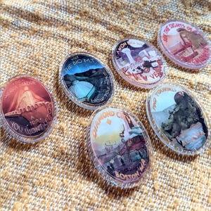 May include: A collection of oval-shaped clear acrylic pins featuring colorful illustrations and text. Each pin showcases a different location, including "The Sierra Madre," "The Big MT," and "Diamond City." The pins are arranged on a textured, tan surface.