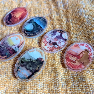 May include: Oval-shaped, clear acrylic pins with colorful illustrations and text. The pins feature various designs, including landscapes and text like "Visit the Big MT" and "See the sights of Emojave". They are arranged on a textured, yellow surface.
