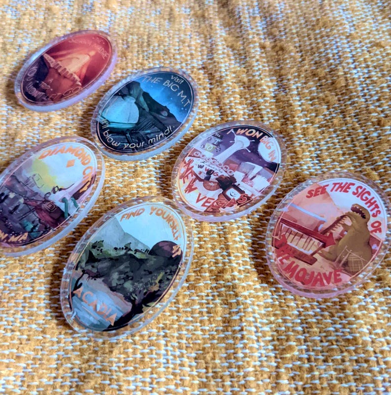 May include: Oval-shaped, clear acrylic pins with colorful illustrations and text. The pins feature various designs, including landscapes and text like "Visit the Big MT" and "See the sights of Emojave". They are arranged on a textured, yellow surface.