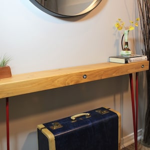 May include: A wooden console table with a natural wood finish and red hairpin legs. The table is decorated with two small potted plants, a book, and a vase with yellow flowers. The table is 122 cm long and 30 cm wide.