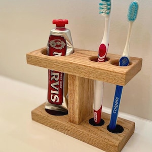 May include: A wooden toothbrush holder with a tube of toothpaste and two toothbrushes. The toothpaste tube is red and silver with the text "N RVIS". One toothbrush is red and white, and the other is blue and white. The holder is made of light-coloured wood.