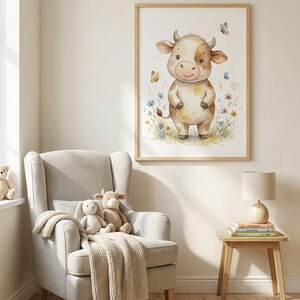 Peut inclure: Une impression encadr&eacute;e &agrave; l'aquarelle d'une vache de dessin anim&eacute; avec un pelage brun clair et blanc, debout dans un champ de fleurs. L'&oelig;uvre est dans un cadre en bois clair et est expos&eacute;e au-dessus d'un fauteuil gris.