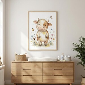 Peut inclure: Une impression encadr&eacute;e &agrave; l'aquarelle d'une vache de dessin anim&eacute; avec des marques marron et blanches, debout dans un champ de fleurs sauvages. Des papillons voltigent autour de la vache. L'impression est affich&eacute;e au-dessus d'une commode en bois.