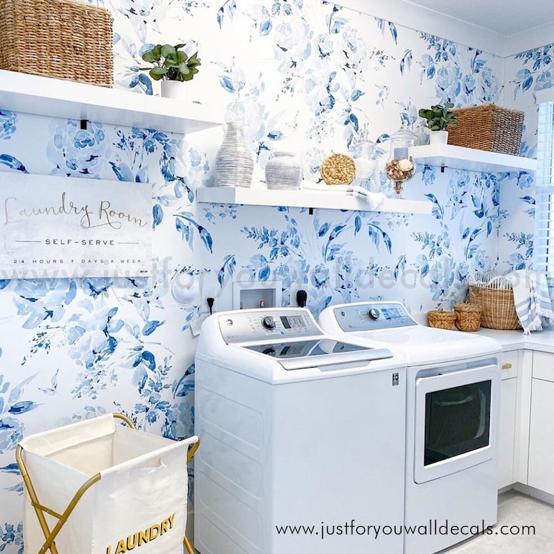 May include: A laundry room with white appliances, shelves, and a floral wallpaper in shades of blue. A white laundry basket with gold accents sits in the foreground. The sign reads "Laundry Room Self-Serve 24 Hours 7 Days a Week."