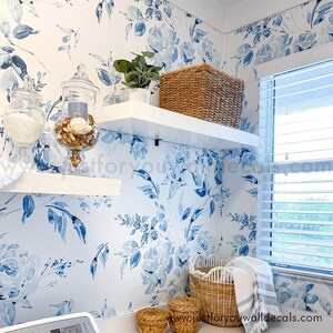 May include: A laundry room with blue floral wallpaper and white shelves. A woven basket and a small potted plant sit on the shelves. Glass jars with decorative items and woven baskets are on the lower shelves. A window with blinds is visible.
