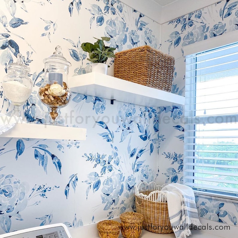 May include: A laundry room with blue floral wallpaper and white shelves. A woven basket and a small potted plant sit on the shelves. Glass jars with decorative items and woven baskets are on the lower shelves. A window with blinds is visible.