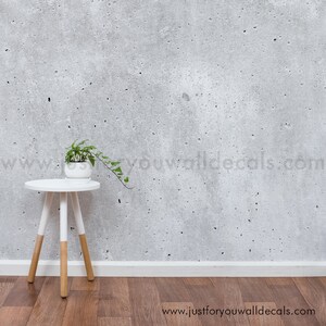 May include: A white side table with wooden legs sits on a wood floor in front of a gray concrete wall. A small potted plant with green leaves sits on the table.