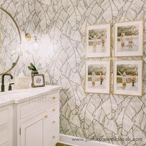 May include: A bathroom featuring a white vanity, gold fixtures, and a circular mirror. The walls are adorned with black and white banana leaf wallpaper. Four framed photographs are displayed, with a small framed photo on the vanity.