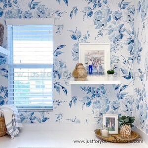 May include: A laundry room with white walls adorned with a blue floral wallpaper. A white shelf holds a framed photo, a woven basket, and a small plant. A window with blinds is visible. The website address "www.justforyouwalldecals.com" is at the bottom.