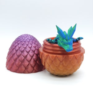 May include: A 3D-printed dragon emerging from a decorative egg. The egg is split open, revealing a small, colorful dragon with blue and green accents. The egg and container are shades of orange and purple.