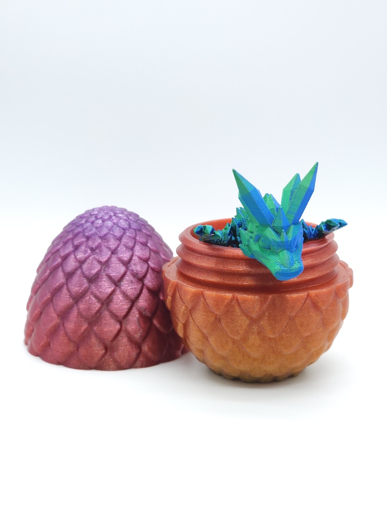 May include: A 3D-printed dragon emerging from a decorative egg. The egg is split open, revealing a small, colorful dragon with blue and green accents. The egg and container are shades of orange and purple.