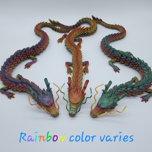 May include: Three rainbow-coloured dragon figurines are displayed against a white background. The text "Rainbow colour varies" is at the bottom. Each dragon has a detailed, textured design.