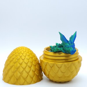 May include: A golden, egg-shaped container with a textured, scale-like pattern. The container is open, revealing a small, multi-colored dragon figurine inside. The lid is placed next to the container. The dragon is blue and green.