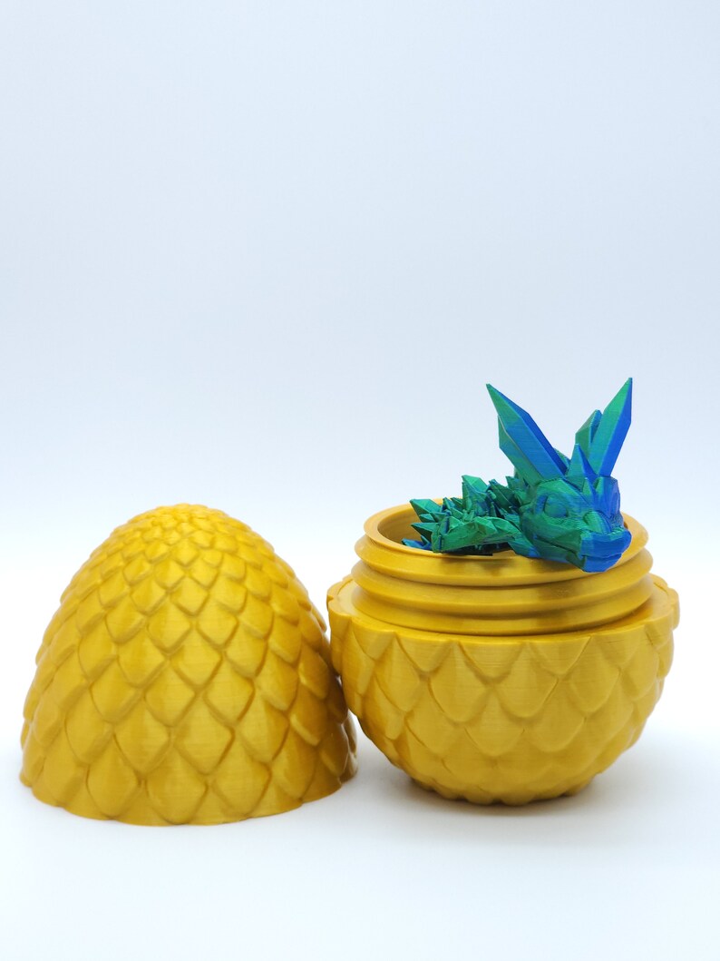 May include: A golden, egg-shaped container with a textured, scale-like pattern. The container is open, revealing a small, multi-colored dragon figurine inside. The lid is placed next to the container. The dragon is blue and green.