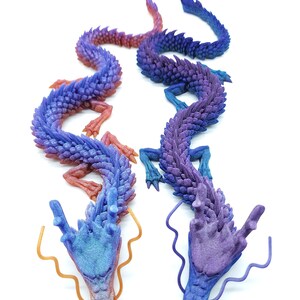 May include: Two detailed, articulated dragon sculptures in shades of blue, purple, and red. The dragons have intricate scale patterns and flowing forms, with golden-coloured tendrils extending from their heads. The background is plain white.