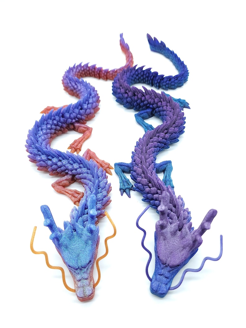 May include: Two detailed, articulated dragon sculptures in shades of blue, purple, and red. The dragons have intricate scale patterns and flowing forms, with golden-coloured tendrils extending from their heads. The background is plain white.