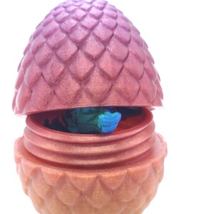 May include: A decorative dragon egg, split open to reveal a colorful interior. The egg is a gradient of pink and orange, with a textured, scale-like pattern. The interior shows blue and green colors.