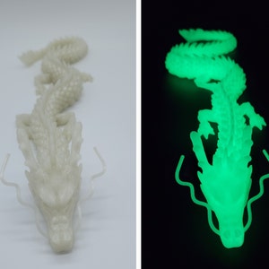 May include: A white, articulated dragon figurine. The dragon is shown in two images, one in daylight and the other glowing bright green in the dark. The dragon has a detailed, textured body and a detailed head with long, curved whiskers.