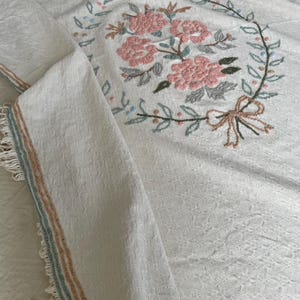 May include: Cream-coloured embroidered floral throw blanket. The blanket features a large floral wreath with pink flowers, green leaves, and a tan bow. The edges have a fringe and stripes in shades of blue and tan.