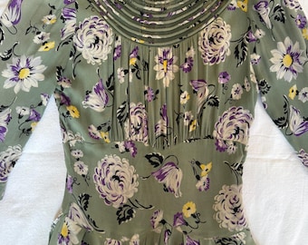 Puede incluir: Vestido vintage verde salvia con un estampado floral de flores blancas, moradas y amarillas. El vestido tiene mangas largas, un cuello alto con detalles plisados y una cintura entallada. El vestido está hecho de una tela suave y fluida.