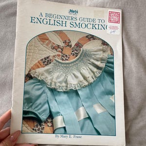 May include: A book titled "A Beginners Guide to English Smocking" by Mary E. Frase. The cover features a blue smocked dress with white lace trim and a white ribbon.